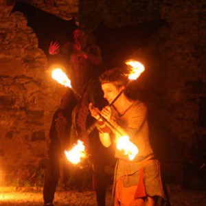 Eine Feuertänzerin mit Feuerstäben und im Hintergrund ein Stelzenläufer im Teufelskostüm bei einer Feuertanztheater Feuershow